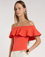 Close-up studio image against a light neutral background, showing an off-the-shoulder coral top with a wide, layered ruffle neckline. 