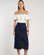 Person standing in a studio against a light neutral background, wearing an off-the-shoulder white top with a wide ruffled neckline paired with a dark blue, textured midi skirt. The skirt features a high waist and a center front slit. 