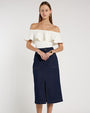 Person standing in a studio against a light neutral background, wearing an off-the-shoulder white top with a wide ruffled neckline paired with a dark blue, textured midi skirt. The skirt features a high waist and a center front slit. 