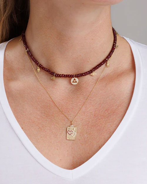 Close-up of two layered necklaces: a dark red beaded choker with gold charms, including an eye symbol and a triangle with an eye, and a longer gold chain with a rectangular pendant engraved with a heart design.