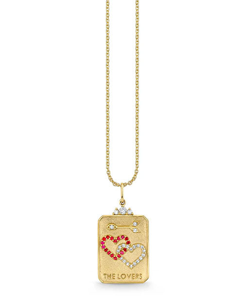 Gold necklace with a rectangular pendant featuring two interlocking hearts—one outlined in red gemstones and the other in white—along with two small fish-like symbols above. The bottom of the pendant is engraved with “THE LOVERS.”