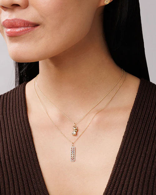 Close-up of two gold necklaces worn over a brown ribbed top. One necklace features a cat-shaped pendant, and the other has a rectangular pendant with colorful gemstones arranged in a pattern.