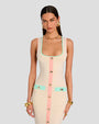 A knee-up view of a brunette model wearing a low-neck, below the knee dress, featuring rectangular blue-pockets and gold buttons, which also appear down the middle on a pink stripe. She is also wearing large gold earrings, gold bracelets and a gold necklace. The background is plain white.