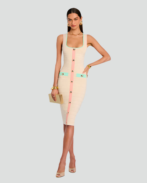 A brunette model with her legs crossed and her hand on her hip wearing a low-neck, below the knee dress, featuring rectangular blue-pockets and gold buttons, which also appear down the middle on a pink stripe. She is also wearing large gold earrings, a gold clutch, gold heels, and a gold bracelet. The background is plain white.