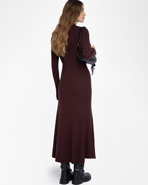 Back view of a person wearing a long, ribbed, dark burgundy knit dress with long sleeves and a flared hem. The dress fits closely through the torso and hips, then widens slightly toward the bottom. The person is carrying a black handbag with a short strap over one shoulder and is wearing chunky black boots with silver hardware. Their long, wavy hair falls down the back.