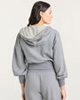 Back view of a gray zip-up hoodie with a drawstring hood and long sleeves. The hoodie features ruffled trim along the edge of the hood and around the waistband. It is paired with matching gray sweatpants that also have ruffled detailing near the pockets. The fabric appears soft and casual, suitable for loungewear.