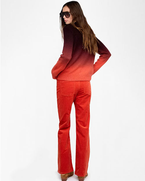 Back view of a person standing against a plain white background wearing a gradient sweater that transitions from dark burgundy at the top to bright red at the bottom. The sweater has long sleeves and a ribbed hem. The outfit includes high-waisted red pants with visible back pockets and brown suede shoes. The person’s hands are placed on the hips, and long wavy hair falls down the back.