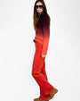 A person is shown in a side view against a plain white background wearing a gradient sweater that transitions from dark burgundy at the top to bright red at the bottom. The sweater has long sleeves and a round neckline. The outfit includes high-waisted red pants with a black belt and brown suede shoes. The person’s left arm is relaxed by the side, and the hair is long and wavy, falling over the shoulder.