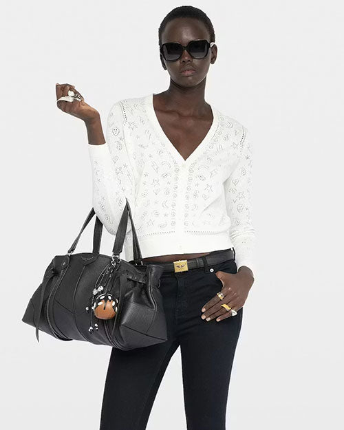 A person wearing a white long‑sleeve V‑neck cardigan with an open‑work embroidered pattern throughout the fabric. The cardigan is slightly cropped and fitted. The individual is also wearing dark, slim‑fit pants and holding a large black leather handbag with multiple straps and decorative keychains attached. The background is plain and light-colored.