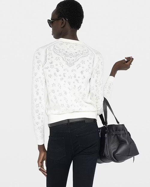 Back view of a person wearing a white long‑sleeve V‑neck cardigan with an open‑work embroidered pattern throughout the fabric. The cardigan is slightly cropped and fitted. The individual is also wearing dark, slim‑fit pants and holding a large black leather handbag. The background is plain and light-colored.