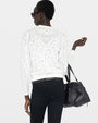 Back view of a person wearing a white long‑sleeve V‑neck cardigan with an open‑work embroidered pattern throughout the fabric. The cardigan is slightly cropped and fitted. The individual is also wearing dark, slim‑fit pants and holding a large black leather handbag. The background is plain and light-colored.