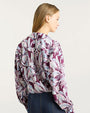 Back view of a long-sleeve blouse featuring a bold, abstract feather-like pattern in shades of purple, lavender, and light blue. The blouse has a relaxed fit and is paired with high-waisted dark denim jeans with visible seams.