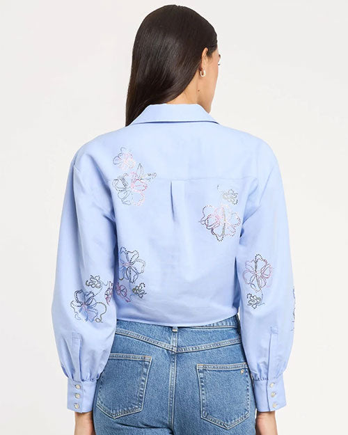 Back view of a light blue long-sleeve blouse with a pointed collar and delicate embroidered floral motifs across the back and sleeves, finished with buttoned cuffs, paired with mid-rise blue jeans, photographed against a light studio background.