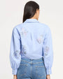 Back view of a light blue long-sleeve blouse with a pointed collar and delicate embroidered floral motifs across the back and sleeves, finished with buttoned cuffs, paired with mid-rise blue jeans, photographed against a light studio background.