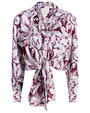 Front view of a long-sleeve button-up blouse with a bold, abstract feather-like pattern in shades of purple, lavender, and light blue. The blouse features a pointed collar and a tie-front detail at the waist.