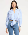 Front view of a light blue button-down blouse with delicate embroidered floral motifs, long sleeves with gathered cuffs, and a tie-front hem, paired with mid-rise blue jeans, photographed against a light studio background.