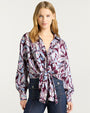 Person wearing a long-sleeve button-up blouse with a bold, abstract feather-like pattern in shades of purple, lavender, and light blue. The blouse is tied in a knot at the waist, paired with high-waisted dark denim jeans featuring prominent front seams and decorative buttons.