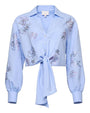 Front view of a light blue cropped button-down blouse featuring delicate embroidered floral motifs, a pointed collar, long sleeves with gathered cuffs, and a tie-front hem, displayed against a white background.