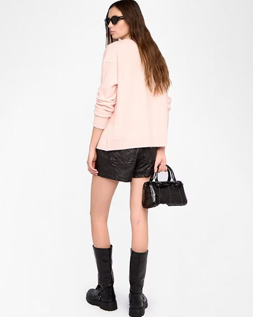  A person is standing against a plain white background, facing slightly away from the camera. The outfit includes a light pink long-sleeve cardigan, black textured shorts, and knee-high black leather boots with buckles. The person is holding a small black handbag with silver stud detailing in the left hand. The hair is long and straight, falling down the back.