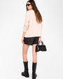  A person is standing against a plain white background, facing slightly away from the camera. The outfit includes a light pink long-sleeve cardigan, black textured shorts, and knee-high black leather boots with buckles. The person is holding a small black handbag with silver stud detailing in the left hand. The hair is long and straight, falling down the back.