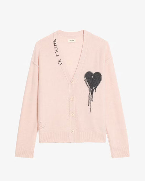A light pink buttoned cardigan displayed against a plain background. The cardigan features a deep V-neckline with beige buttons down the front. On the left chest area, there is a large black heart graphic with a dripping effect. Near the right shoulder, the text “JE T’AIME” is printed vertically in black. The fabric appears soft and knitted, with ribbed cuffs and hem.