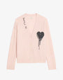 A light pink buttoned cardigan displayed against a plain background. The cardigan features a deep V-neckline with beige buttons down the front. On the left chest area, there is a large black heart graphic with a dripping effect. Near the right shoulder, the text “JE T’AIME” is printed vertically in black. The fabric appears soft and knitted, with ribbed cuffs and hem.