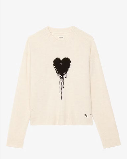 A cream-colored long-sleeve sweatshirt displayed against a plain background. The sweatshirt features a large black heart graphic with a dripping paint effect in the center of the chest. There is also small black text near the bottom right hem that reads “JE T’AIME.” The design is simple and modern with ribbed cuffs and hem.