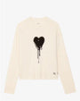 A cream-colored long-sleeve sweatshirt displayed against a plain background. The sweatshirt features a large black heart graphic with a dripping paint effect in the center of the chest. There is also small black text near the bottom right hem that reads “JE T’AIME.” The design is simple and modern with ribbed cuffs and hem.