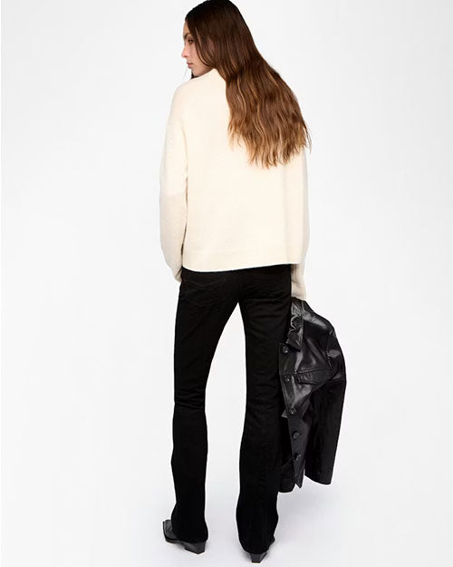 A person is standing against a plain white background, shown from the back, wearing a cream-colored long-sleeve sweatshirt and black flared pants. The outfit is paired with black shoes, and the person is holding a black leather jacket in the left hand. The hair is long and wavy, cascading down the back.