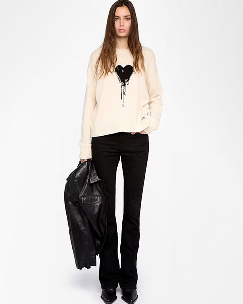 A person is standing against a plain white background wearing a cream-colored long-sleeve sweatshirt featuring a large black heart graphic with a dripping effect on the chest and small text detail on the left sleeve. The outfit includes black flared pants and black shoes. The person is holding a black leather jacket in the left hand.