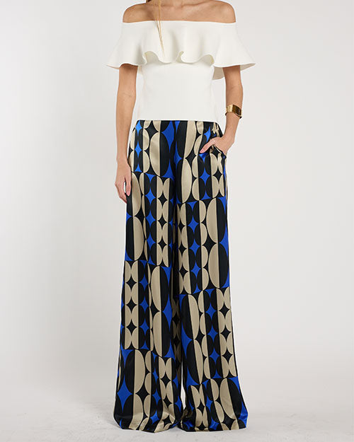 Person standing in a studio against a light neutral background, wearing an off-the-shoulder white top with a layered ruffle neckline paired with wide-leg patterned pants featuring geometric shapes in blue, beige, and black. 