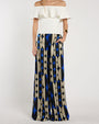 Person standing in a studio against a light neutral background, wearing an off-the-shoulder white top with a layered ruffle neckline paired with wide-leg patterned pants featuring geometric shapes in blue, beige, and black. 