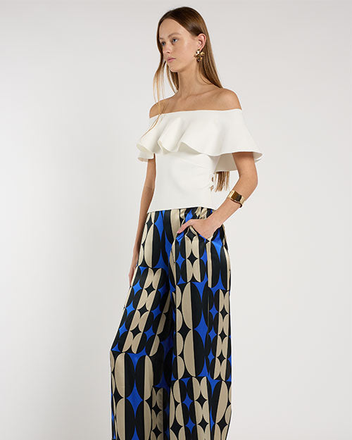 Person standing in a studio against a light neutral background, wearing an off-the-shoulder white top with a layered ruffle neckline paired with wide-leg patterned pants featuring geometric shapes in blue, beige, and black. 