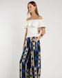 Person standing in a studio against a light neutral background, wearing an off-the-shoulder white top with a layered ruffle neckline paired with wide-leg patterned pants featuring geometric shapes in blue, beige, and black. 