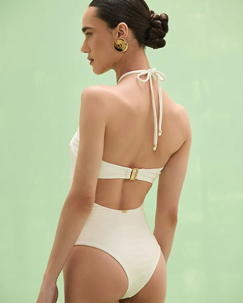 Back studio view of a cream-colored halter-neck bodysuit with a fitted silhouette, featuring a gold ring detail at the neckline and a smooth, high-cut leg opening, photographed against a light green background.