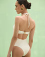 Back studio view of a cream-colored halter-neck bodysuit with a fitted silhouette, featuring a gold ring detail at the neckline and a smooth, high-cut leg opening, photographed against a light green background.