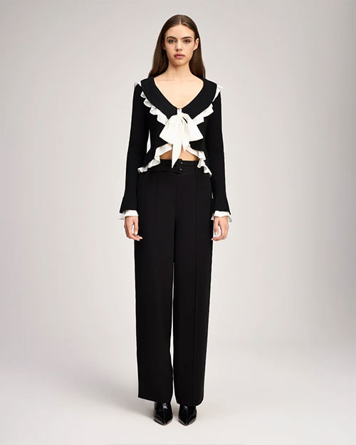 Person standing against a light gray background wearing a black long-sleeve cropped cardigan with white ruffled trim along the neckline, front, hem, and cuffs. The cardigan has a deep V-neck and a large white bow tied at the front, revealing a small section of the midriff. The outfit is paired with high-waisted black wide-leg trousers. 