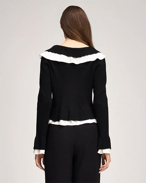 Back view of a person standing against a light gray studio background wearing a fitted black knit cardigan with long sleeves. The cardigan features white ruffled trim across the upper back, along the hem, and at the cuffs, creating a contrast against the black fabric. The garment has a slightly flared peplum-style hem and is paired with high-waisted black wide-leg trousers.