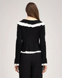 Back view of a person standing against a light gray studio background wearing a fitted black knit cardigan with long sleeves. The cardigan features white ruffled trim across the upper back, along the hem, and at the cuffs, creating a contrast against the black fabric. The garment has a slightly flared peplum-style hem and is paired with high-waisted black wide-leg trousers.