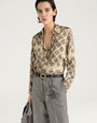 Close-up front view of a cream-colored button-front blouse featuring an allover black floral-inspired print. The long-sleeve blouse has a pointed collar and relaxed fit and is styled tucked into gray plaid high-waisted trousers with a black leather belt and layered necklaces. Shown against a light gray background.