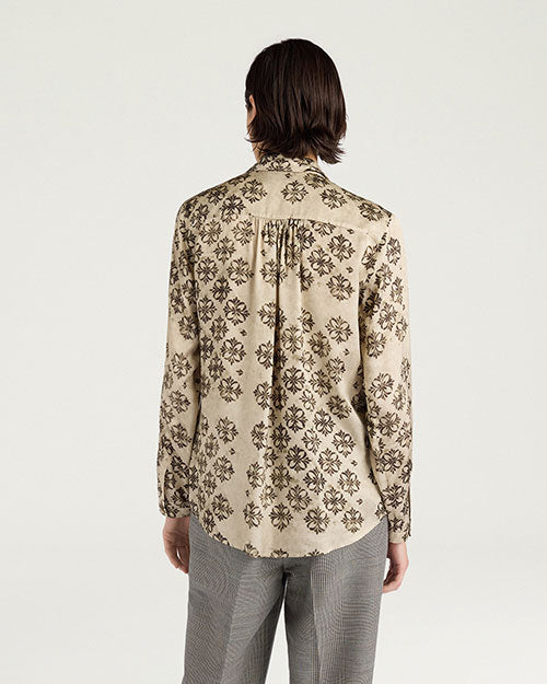 Back view of a cream-colored long-sleeve blouse featuring an allover black floral-inspired print. The blouse has a pointed collar, gathered detailing below the back yoke, buttoned cuffs, and a gently curved hem. Styled with gray plaid trousers and shown against a light gray background.