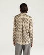 Back view of a cream-colored long-sleeve blouse featuring an allover black floral-inspired print. The blouse has a pointed collar, gathered detailing below the back yoke, buttoned cuffs, and a gently curved hem. Styled with gray plaid trousers and shown against a light gray background.
