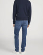 Back view of a person standing in a studio against a light neutral background, wearing a navy crewneck sweater with ribbed cuffs and hem, paired with medium-wash straight-leg jeans and white lace-up sneakers. The image is photographed in a full-length, front-facing pose.