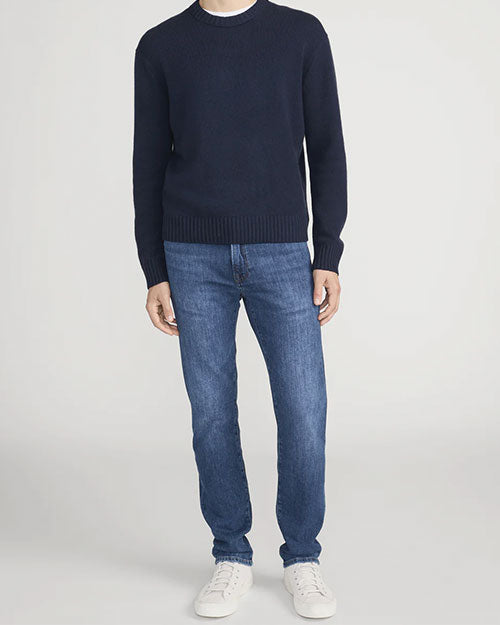 Person standing in a studio against a light neutral background, wearing a navy crewneck sweater with ribbed cuffs and hem, paired with medium-wash straight-leg jeans and white lace-up sneakers. The image is photographed in a full-length, front-facing pose.