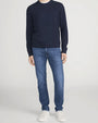 Person standing in a studio against a light neutral background, wearing a navy crewneck sweater with ribbed cuffs and hem, paired with medium-wash straight-leg jeans and white lace-up sneakers. The image is photographed in a full-length, front-facing pose.