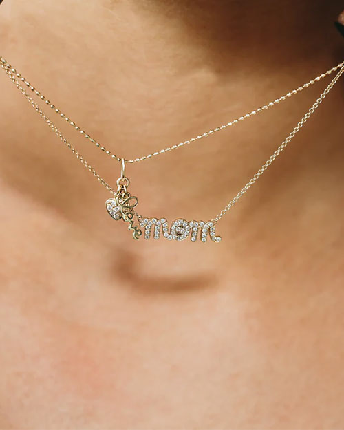 Close-up of a gold necklace with two chains. One chain features a pendant spelling “mom” in cursive letters adorned with sparkling stones, along with a small bee-shaped charm.