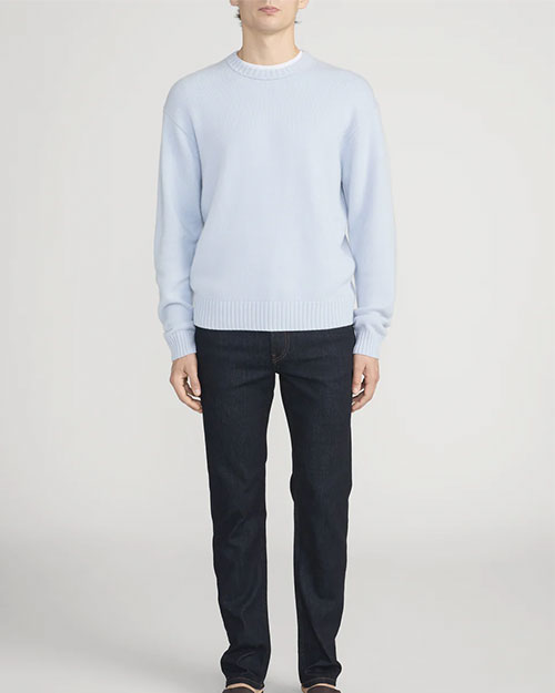 Person standing in a studio against a light neutral background, wearing a light blue crewneck sweater with ribbed neckline, cuffs, and hem, paired with dark-wash straight-leg jeans. The image is photographed in a full-length, front-facing pose.