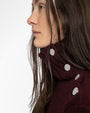 Close-up of a dark burgundy ribbed turtleneck sweater featuring four decorative silver buttons along the side of the collar. The buttons have a textured, sparkling surface, adding a stylish detail to the knit fabric. Part of the wearer’s long, straight hair is visible over the shoulder.