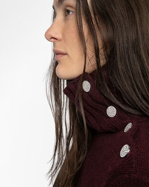 Close-up of a dark burgundy ribbed turtleneck sweater featuring four decorative silver buttons along the side of the collar. The buttons have a textured, sparkling surface, adding a stylish detail to the knit fabric. Part of the wearer’s long, straight hair is visible over the shoulder.