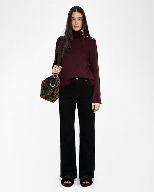 A person wearing a dark burgundy turtleneck sweater with button details on the shoulder, paired with black high-waisted pants and black open-toe sandals. The outfit is accessorized with multiple rings and a leopard-print shoulder bag with a black strap.